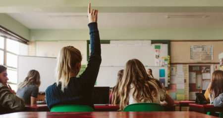 View from behind a classroom of students as one raises her hand.
