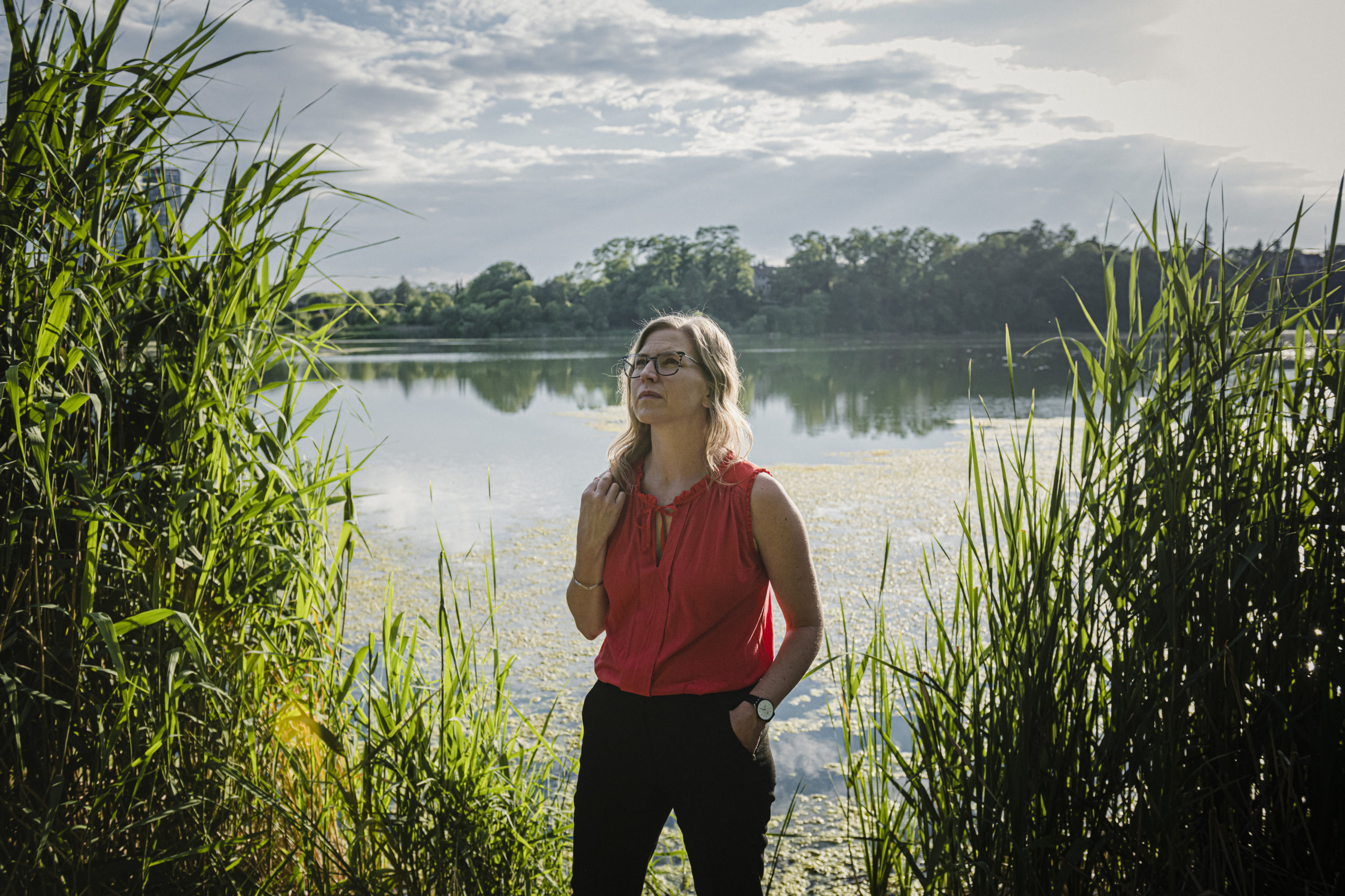 Dayna Nadine Scott stands in front of a pond, framed by tall grasses on a bright but cloudy day.