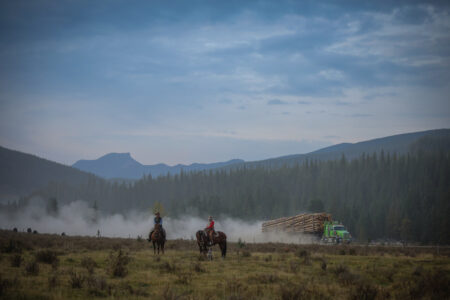 John Smith and Laura Laing ride on horses while log truck passes in the background