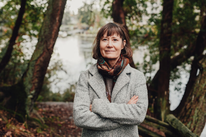 A photo of a woman standing with her arms crossed in a grey jacked in the forest