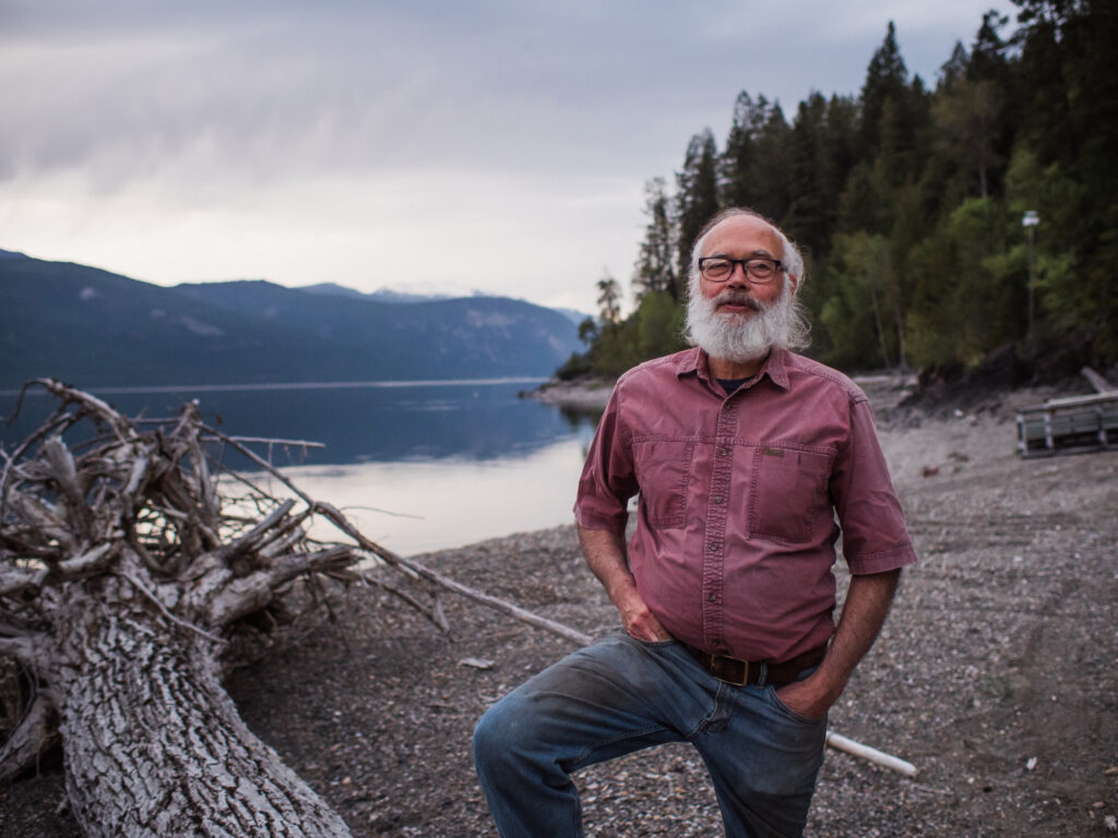 A man with a white beard and a salmon-coloured button-up shirt poses on a rocky lake shore