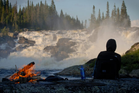 person sits by a fire in front of Muteshekau Shipu
