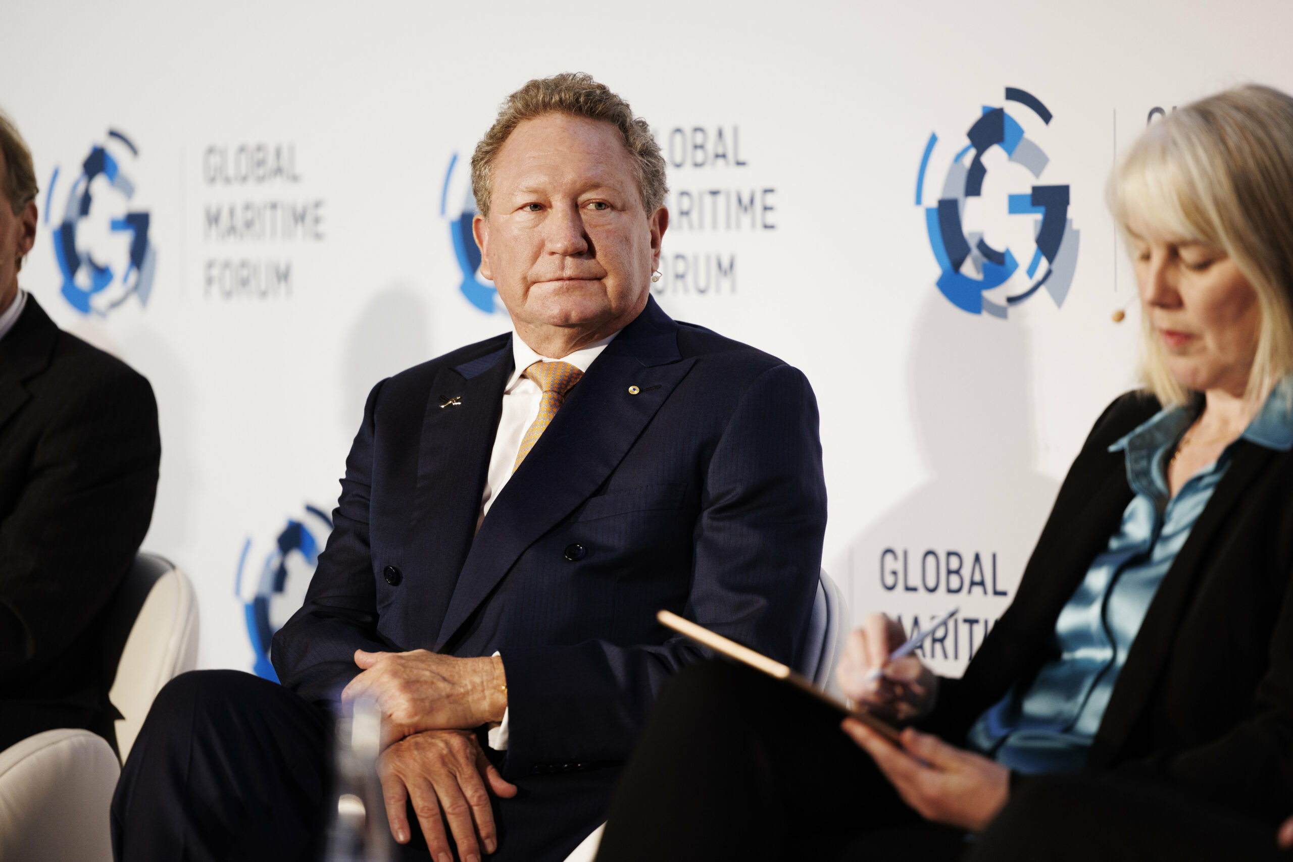 A man in a suit sits in front of a backdrop for an event as a woman with a clipboard sits in the foreground, out of focus