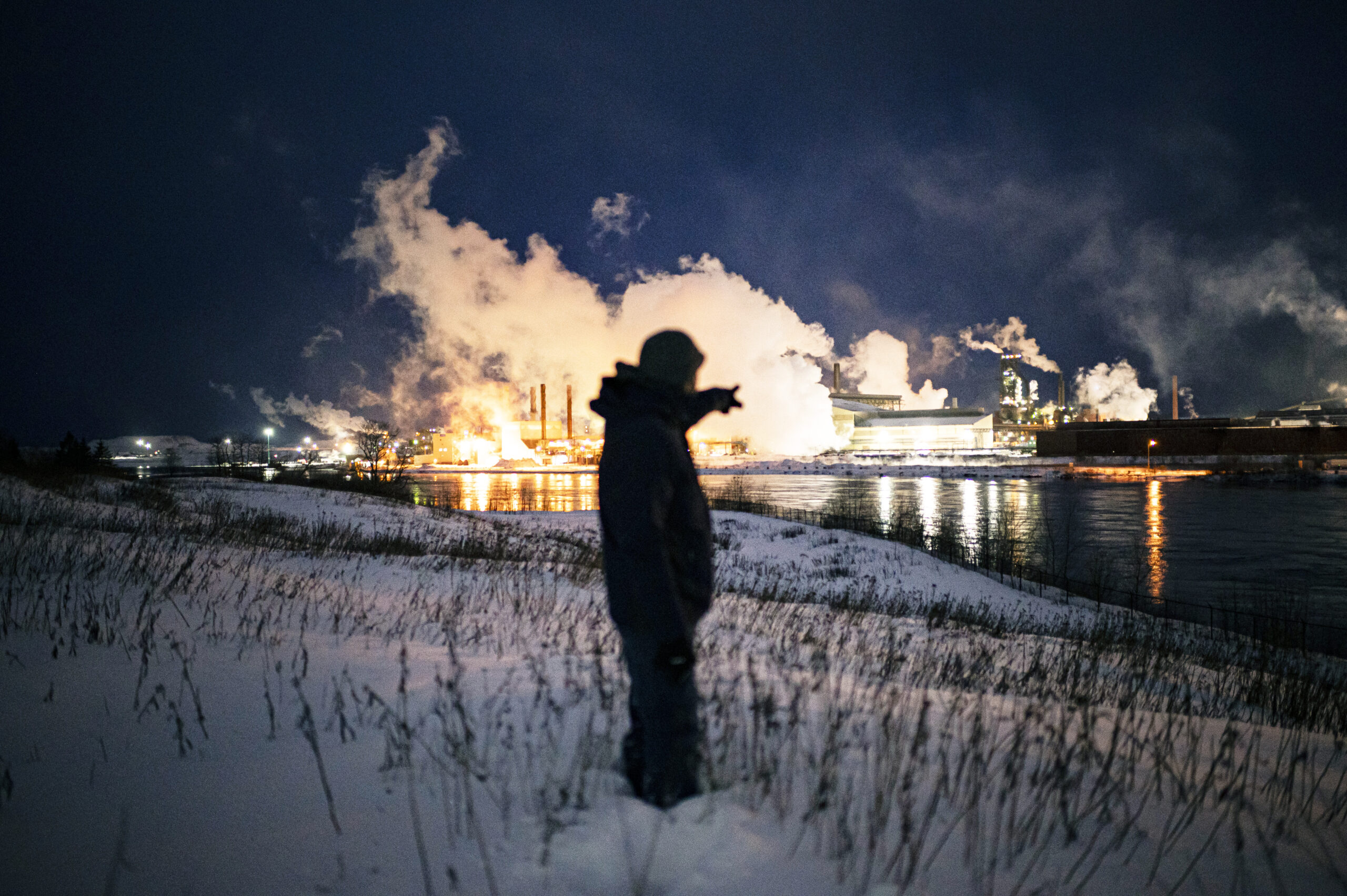 The silhouette of a person standing in snow by a river points to steam rising from the Algoma Steel plant at night. 