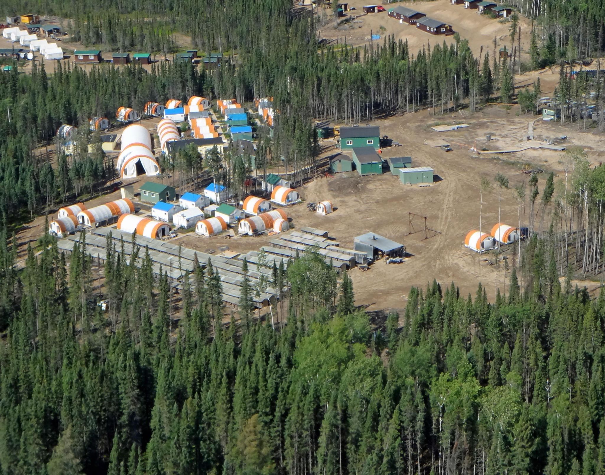 An aerial view of a camp in a clearing in the forest, with buildings and tents with rounded roofs.