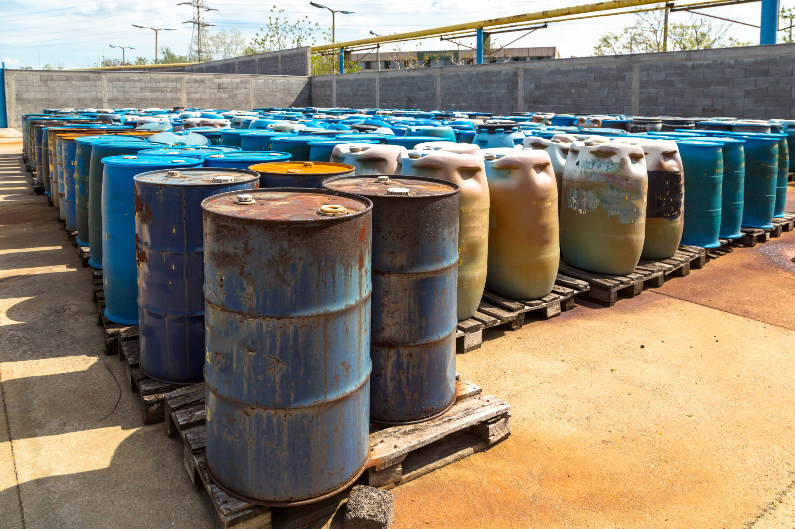A group of dented, rusty barrels holding hazardous waste lined up on wooden pallets. 