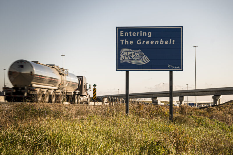 A blue highway sign on the side of a highway reads "Entering the Greenbelt," as a cargo truck passes alongside it