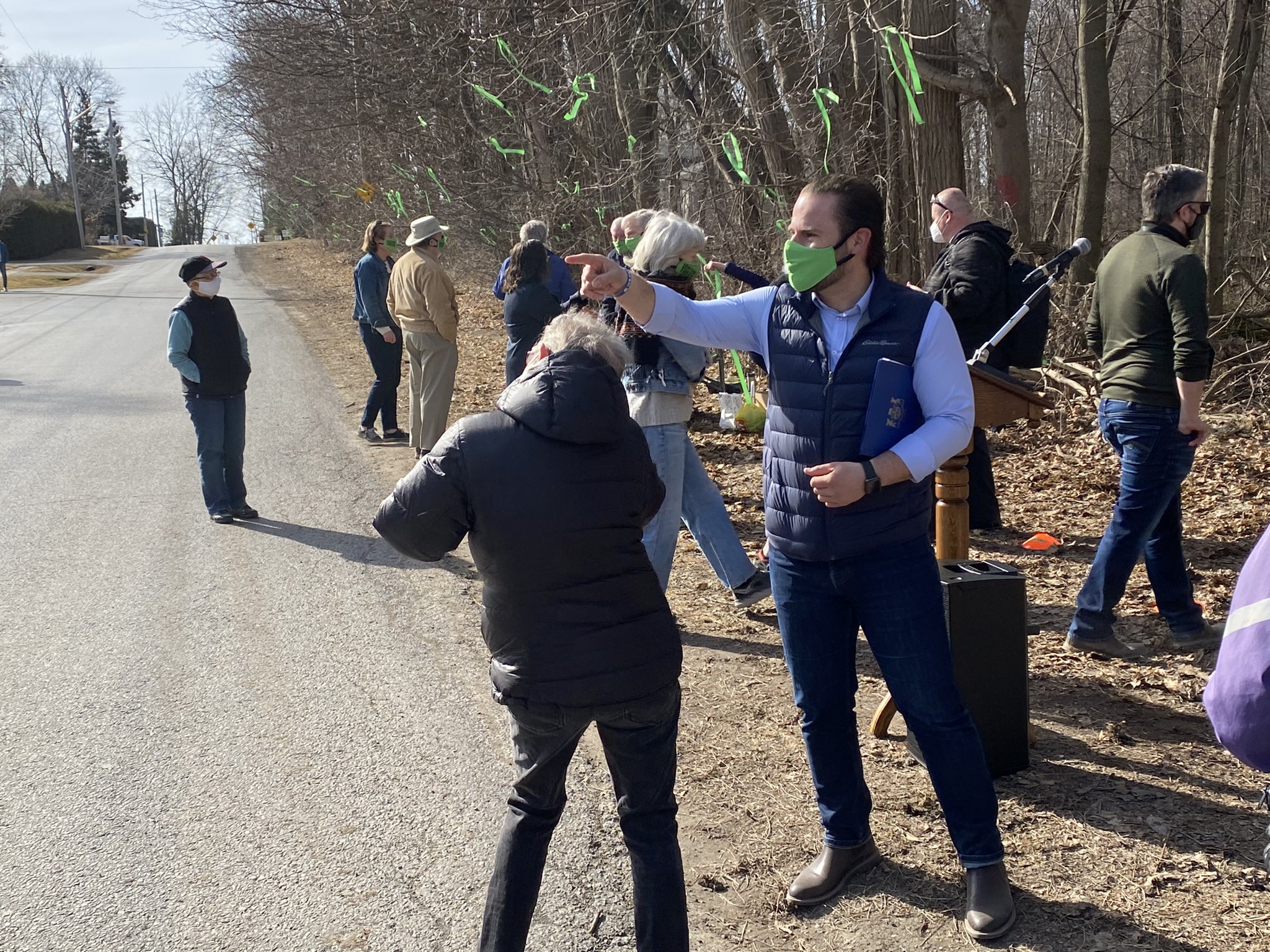 Piccini, wearing jeans and a puffy vest, points off in the distance as he speaks to a person at a protest to save a woodlot. In the background, more attendees are visible in front of trees with green ribbons tied to them.