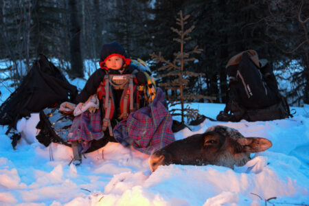 A young child in a carrying sack is perched in the snow in front of a fire