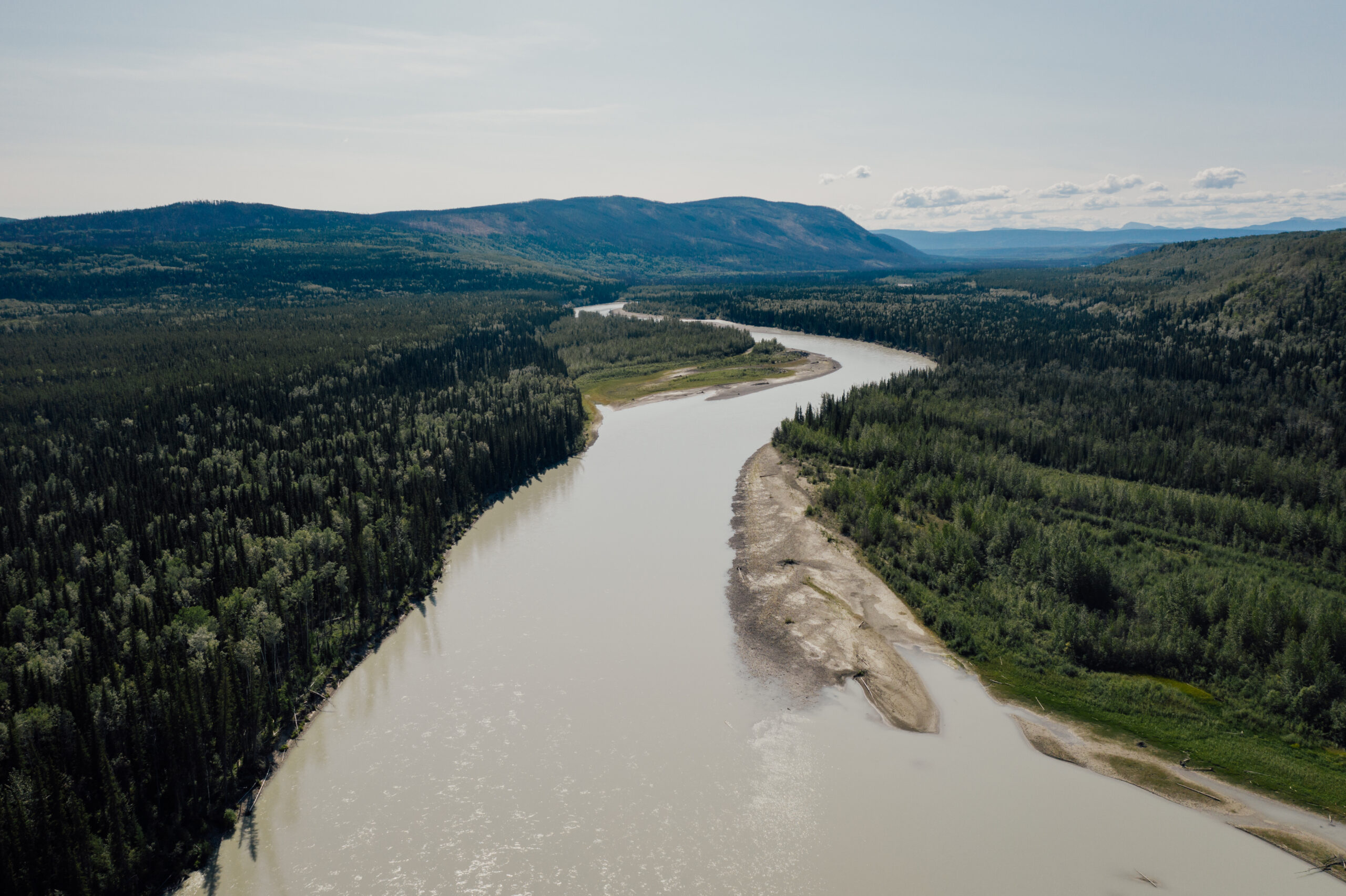 A photo of the Kechika River from the air, running through a forested area, with a mountain in the background. The river runs through the Dene K’éh Kusān Indigenous Protected and Conserved Area