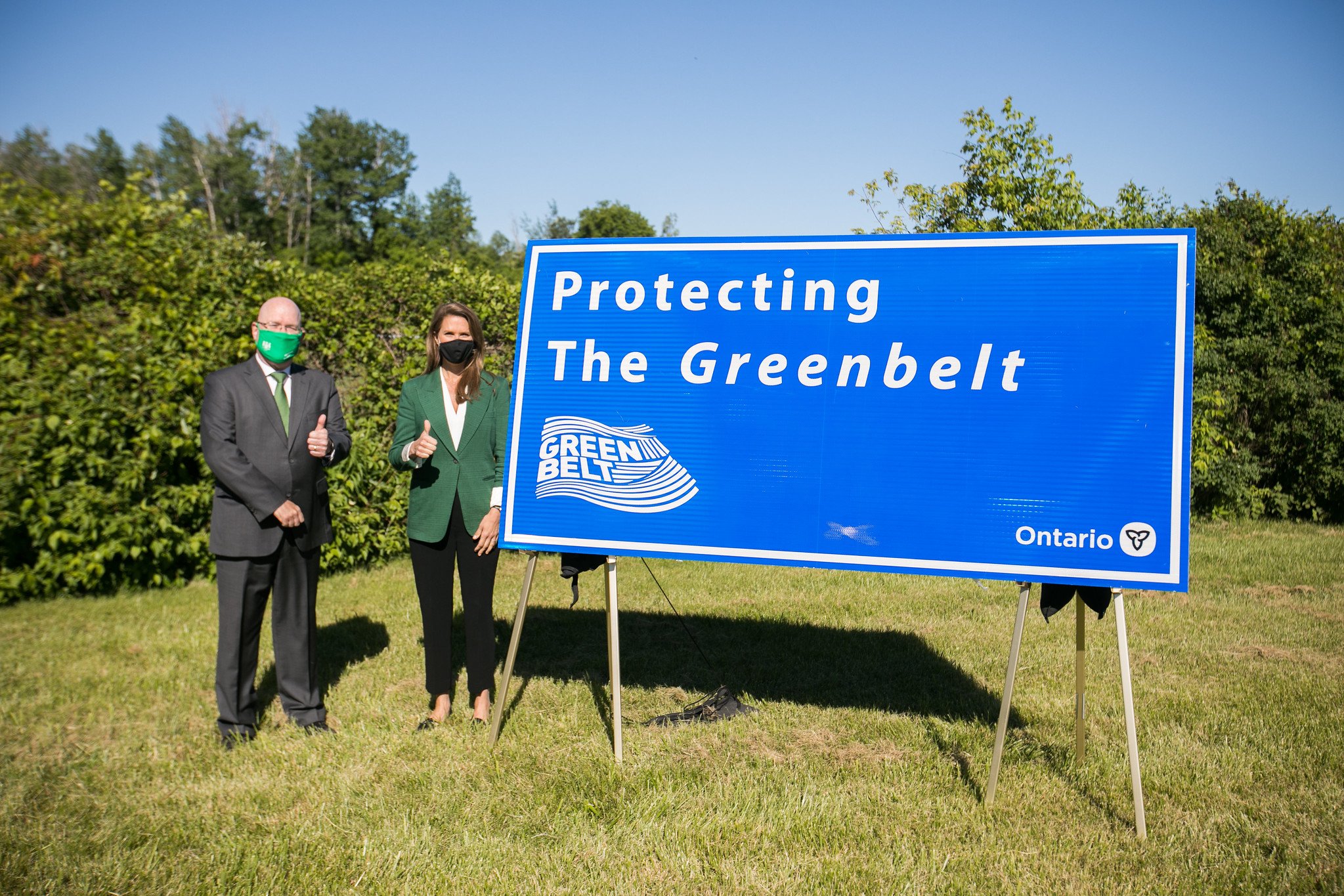 Ontario Municipal Affairs Minister Steve Clark and Transportation Minister Caroline Mulroney, both wearing masks, pose with thumbs up next to a sign that says "Protecting the Greenbelt." Mulroney is spearheading Highway 413.