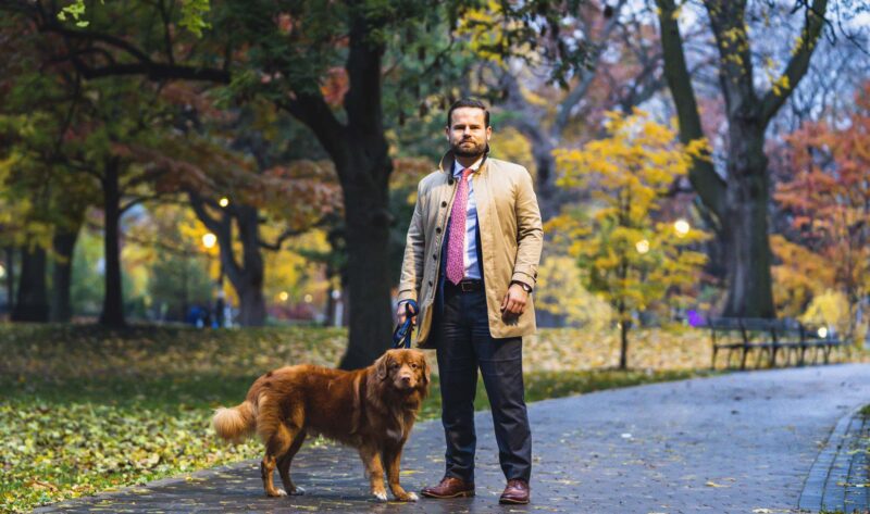 Ontario Environment Minister David Piccini, wearing a jacket and a suit, poses in an autumnal park with his dog Max by his side.