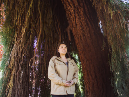 Nuskmata (Jacinda Mack) stands under a tree.