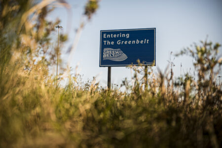 A sign that says "Entering the Greenbelt" stands above grass on a sunny day.