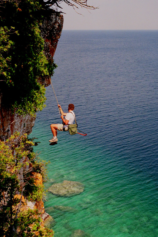 A man wearing shorts and a t-shirt descends down a cliff with a rope and harness, with Lake Huron in the background.
