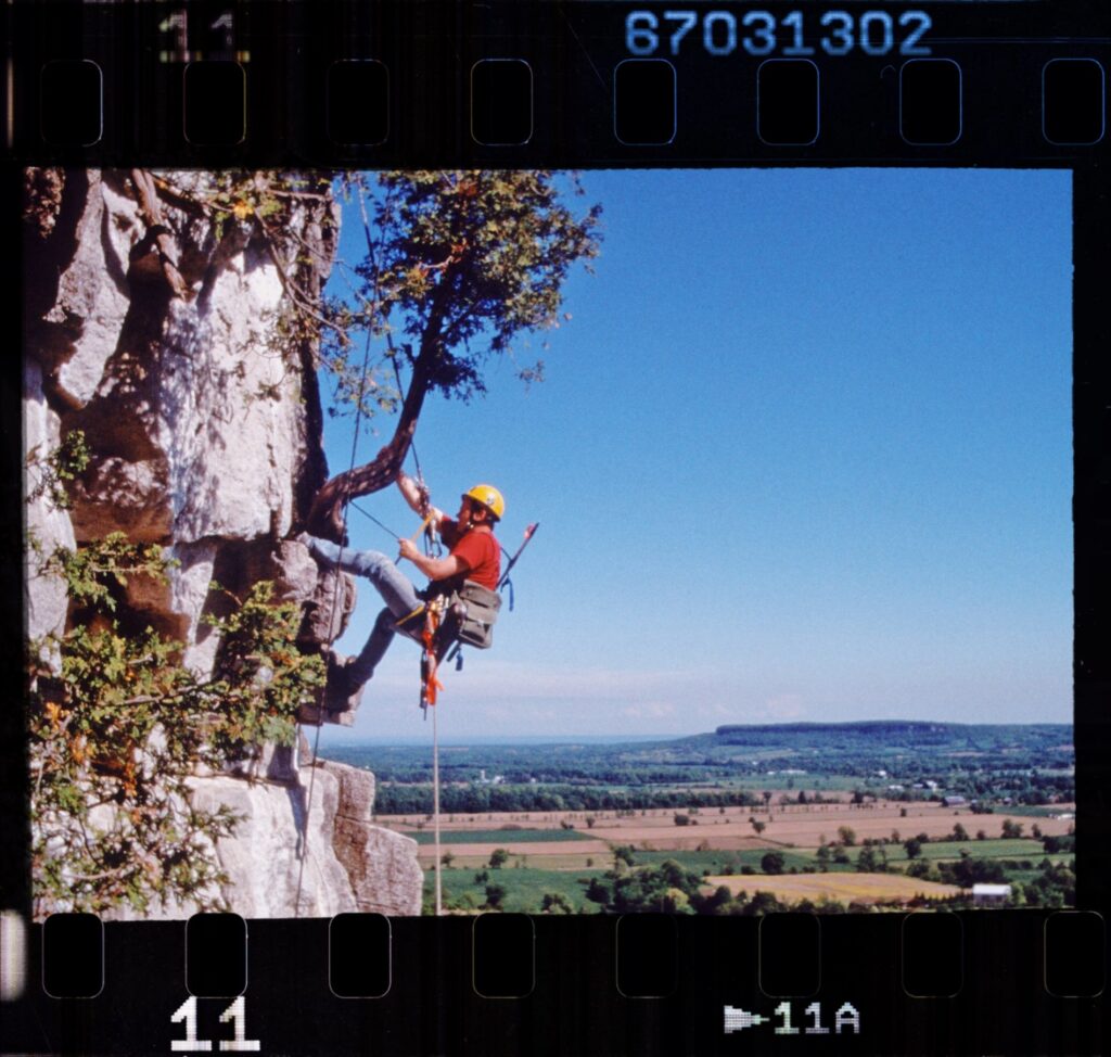 A man hanging from a rope and a climbing harness dangers below a cedar tree growing out of a cliff on the Niagara Escarpment, with farmland and the Niagara Escarpment visible in the background. The photo has a film-style border.