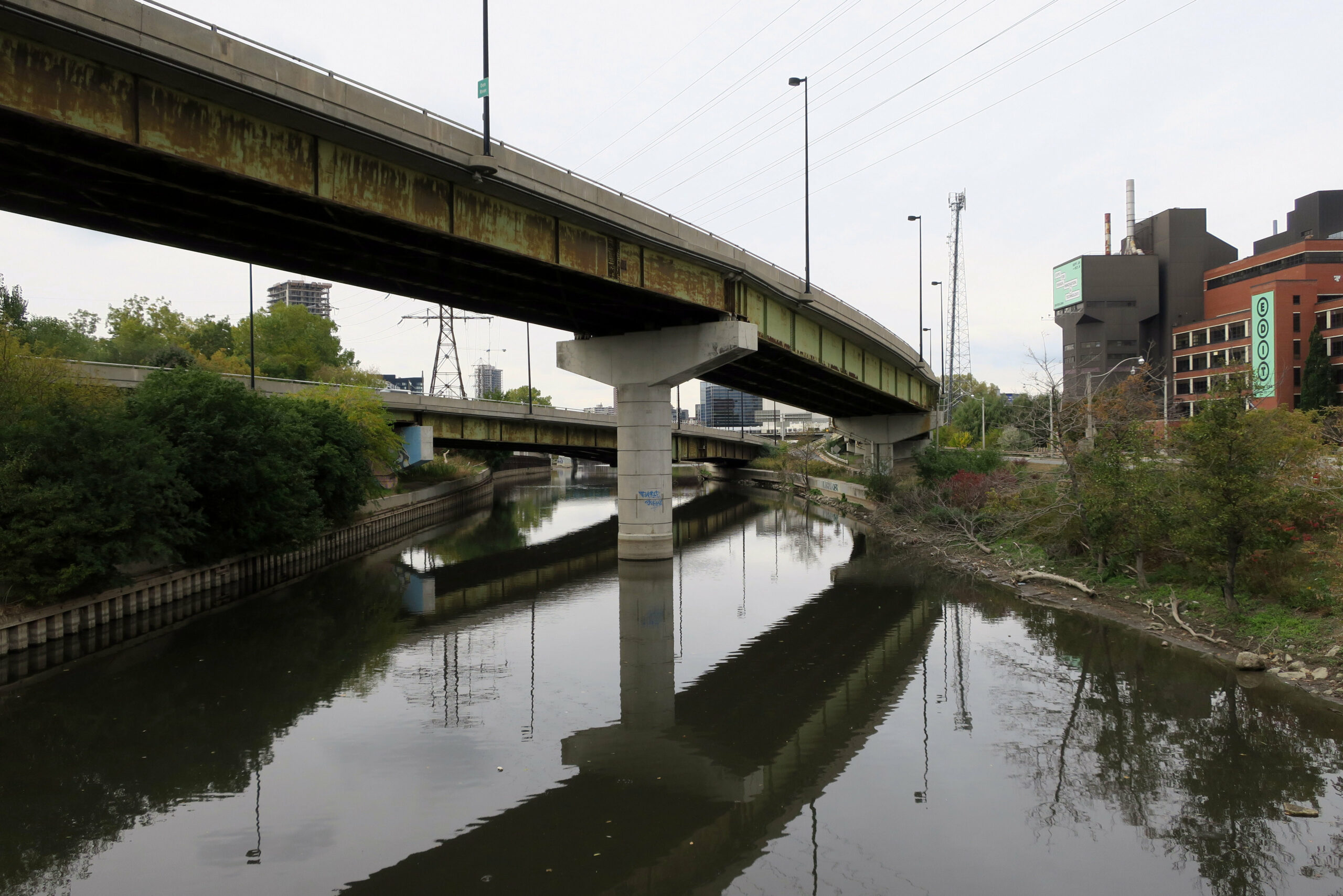 A highway bridge over the muddy water of the Don River in downtown Toronto, with buildings in the background against a grey sky.