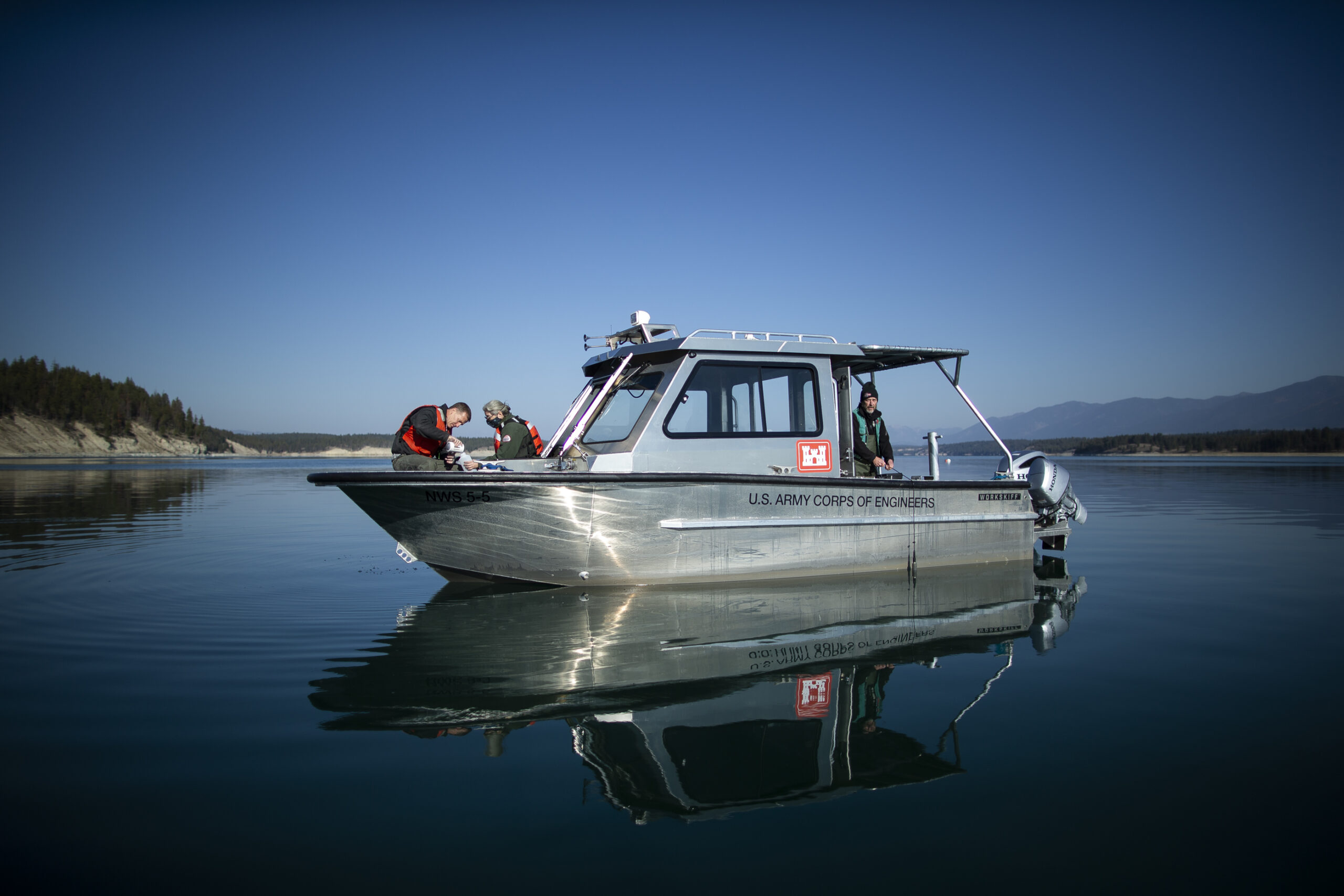 A silver boat sits calmly on deep blue still water in the Koocanusa Reservoir as researchers sample water