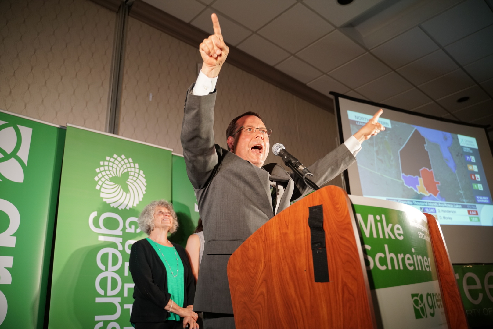 Mike Schreiner stands at a podium, looking animated, with both arms raised, pointing into the air.