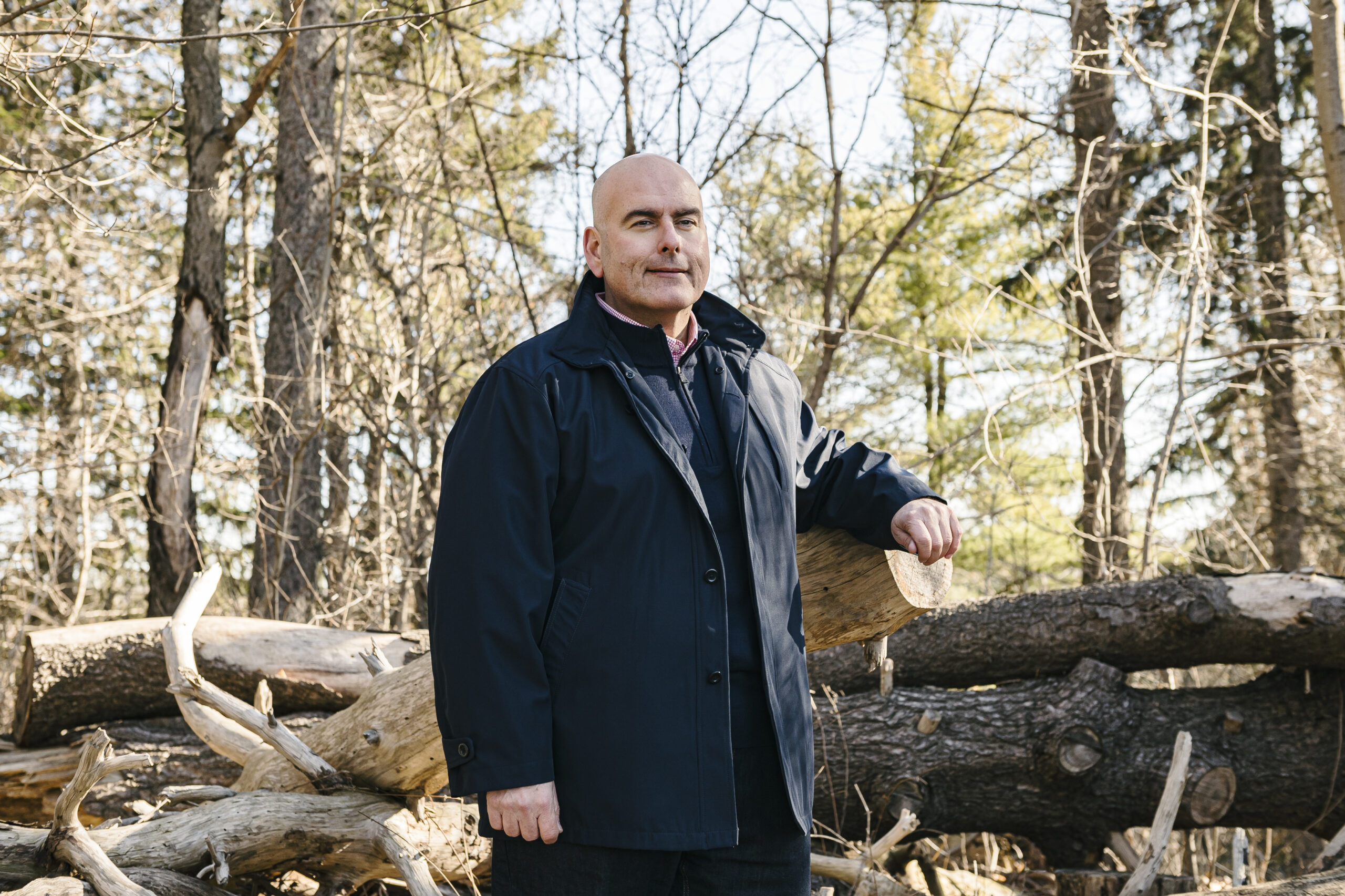 Steven Del Duca leans on a log in a brightly lit forest, smiling slightly.