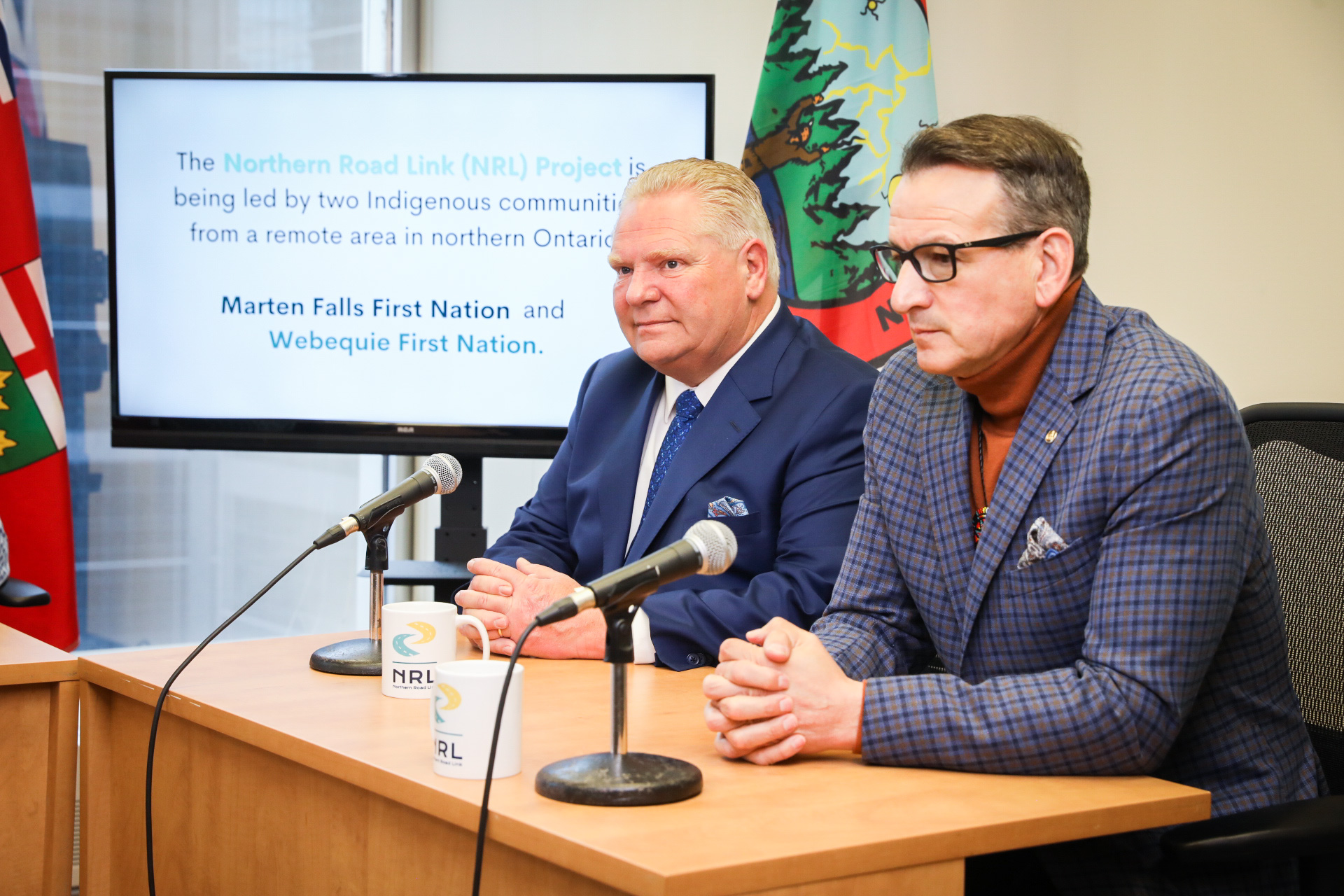 Ontario Premier Doug Ford and Minister of Northern Development, Mines, Natural Resources and Forestry Greg Rickford sit at a table with microphones in front of them, both wearing suits, as they discuss a road for Ontario's Ring of Fire region.