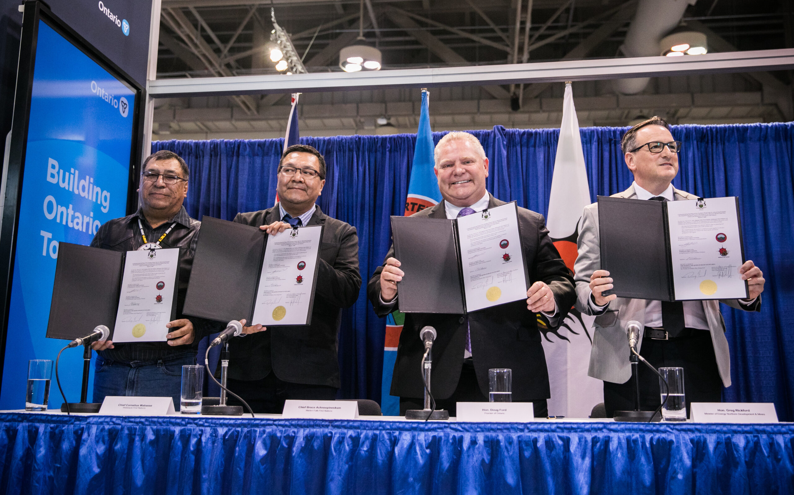 Cornelius Wabasse, Bruce Achneepinkesum, Doug Ford and Greg Rickford hold up signed papers to the camera in front of a blue backdrop.