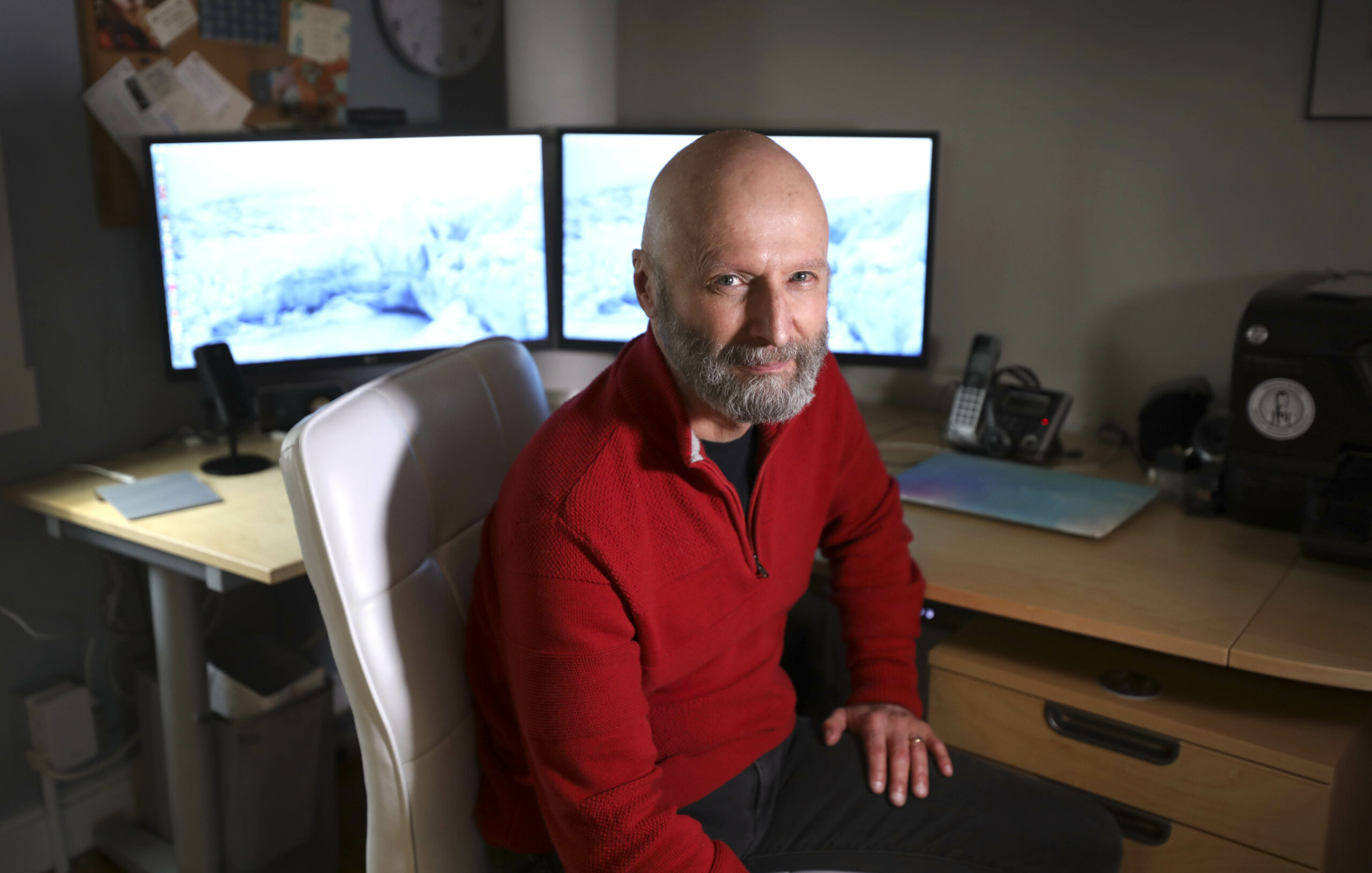 Engineer Curt Hull poses at his home in a red sweater in front of a dual-screen computer.