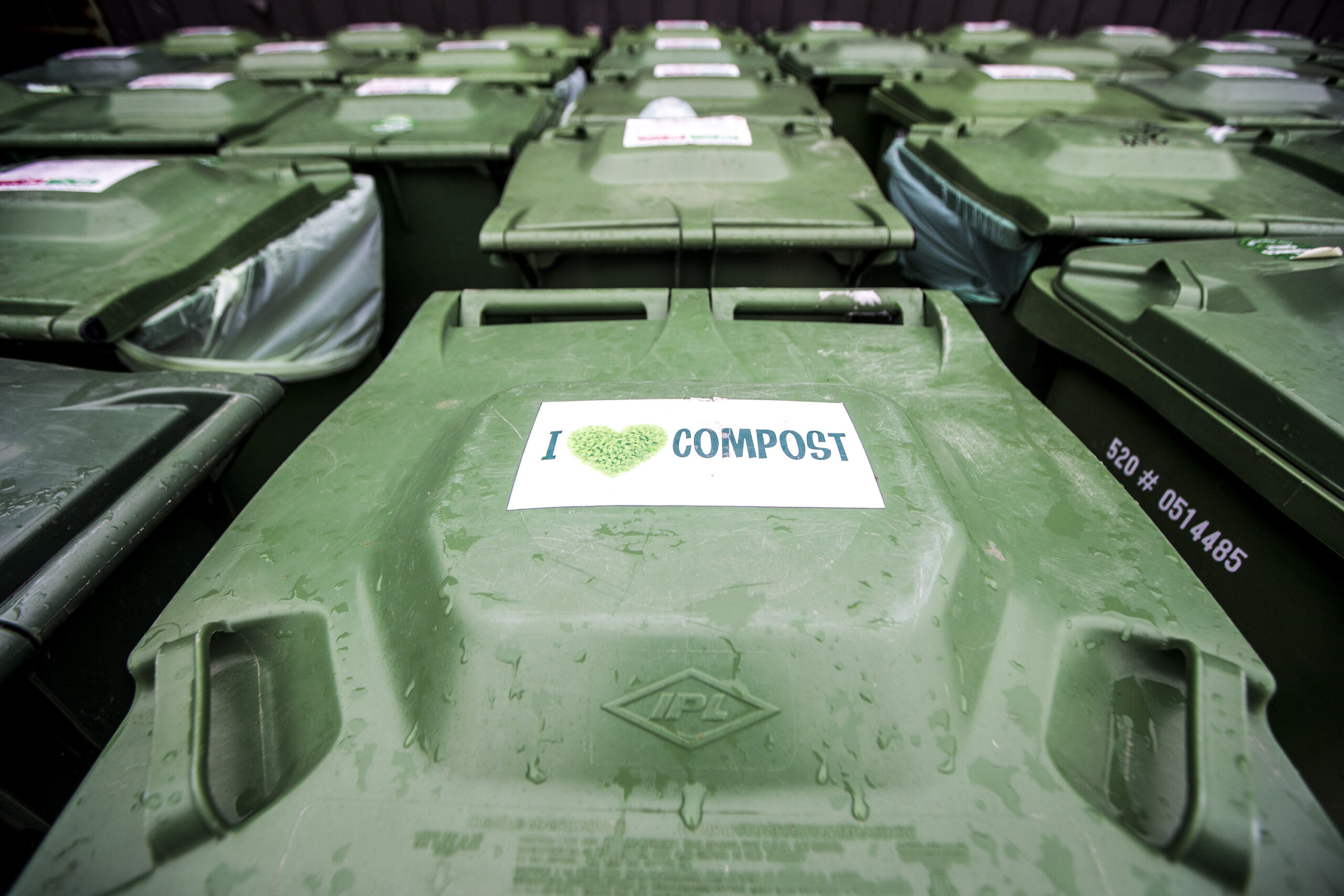 A green compost bin with a sticker reading 'I heart compost' sits among rows of similar bins at Compost Winnipeg headquarters