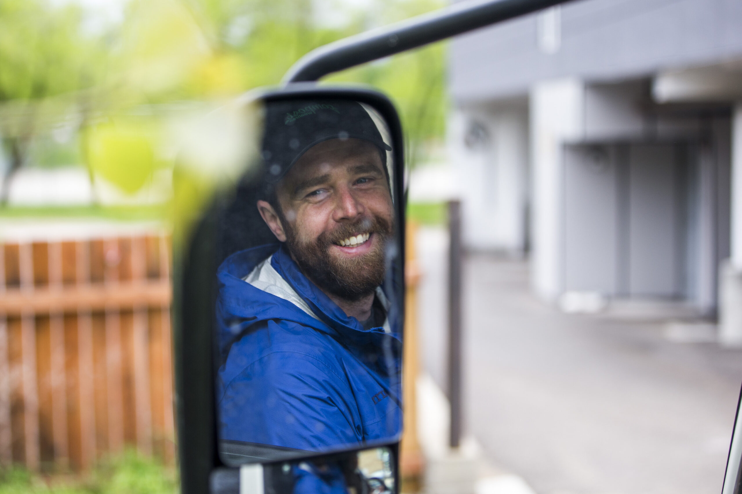 Compost courier Garrett LeBlanc smiles for a portrait in the side mirror of his compost truck during his route