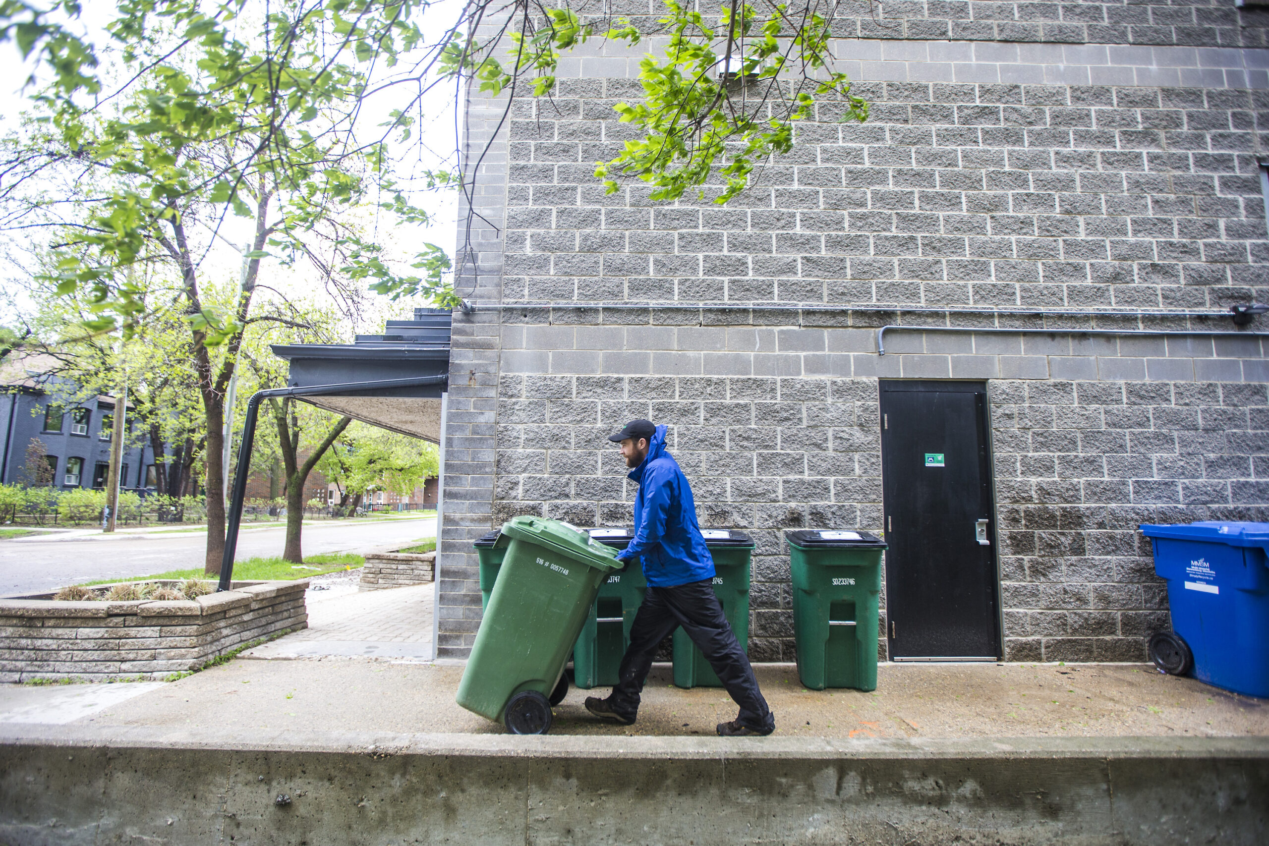 Garrett LeBlanc wheels a green compost bin past a grey brick apartment complex on a rainy day