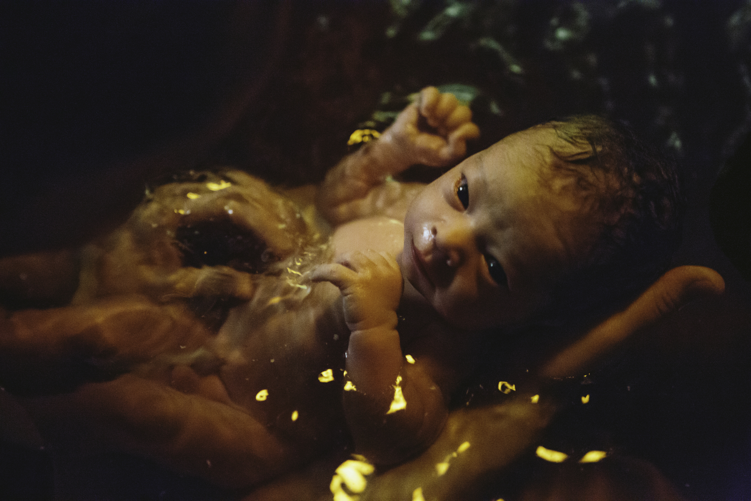 A newborn baby, with umbilical cord still attached, soaks in the water of the birthing pool, eyes gazing up at it's mother