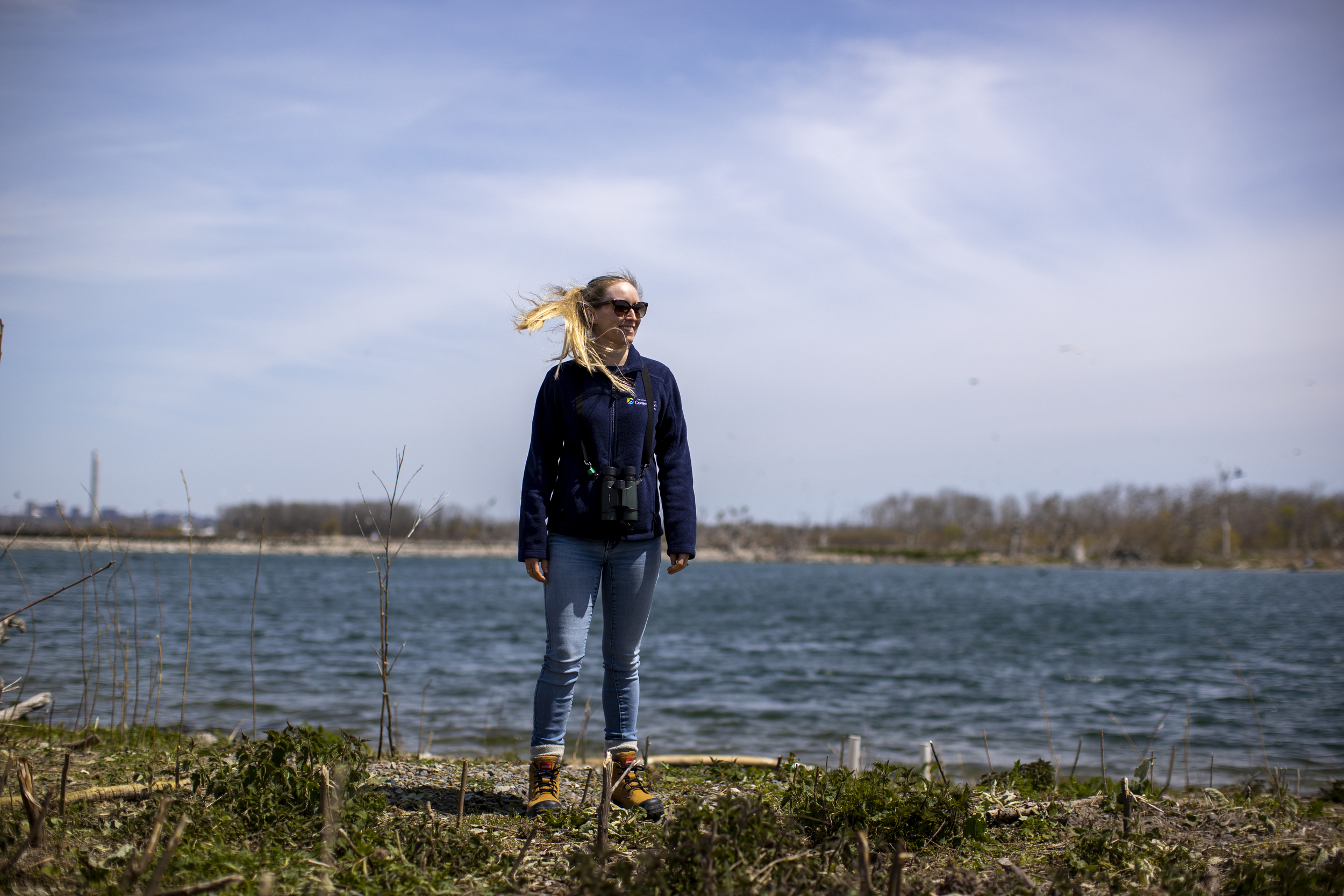 Tommy Thompson Park project manager Andrea Chreston is pictured in sunglasses, jeans, and hiking boots stands on a shoreline in front of water as her blonde ponytail blows in the wind.