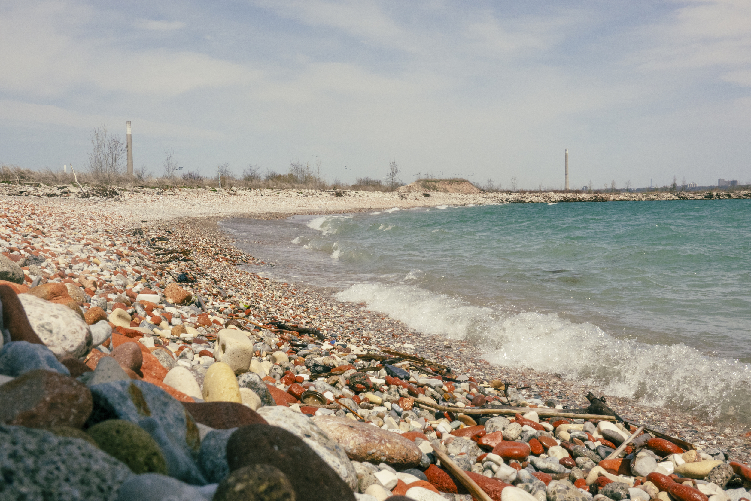 A beach at Tommy Thompson Park with light blue water, covered with gray and brick-red stones.