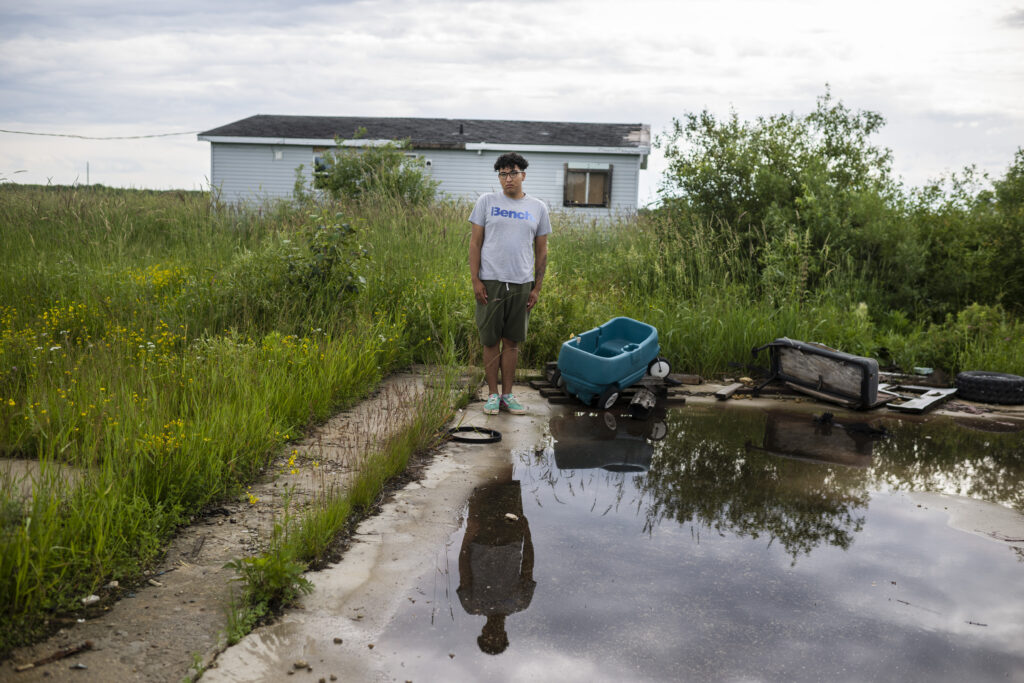 A young man in a grey shirt stands behind a large puddle. A single-story grey home is visible in the background.