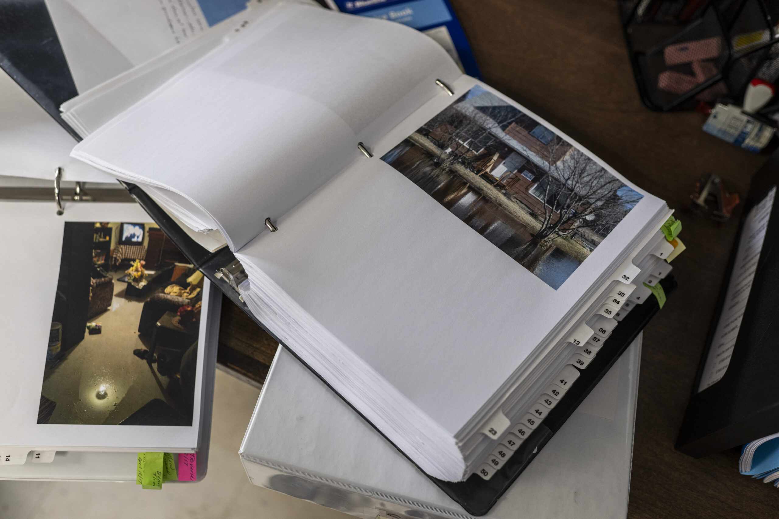 A thick three-ring binder flipped open to a page showing a flood damaged home in Peguis First Nation sits atop a pile of flood papa