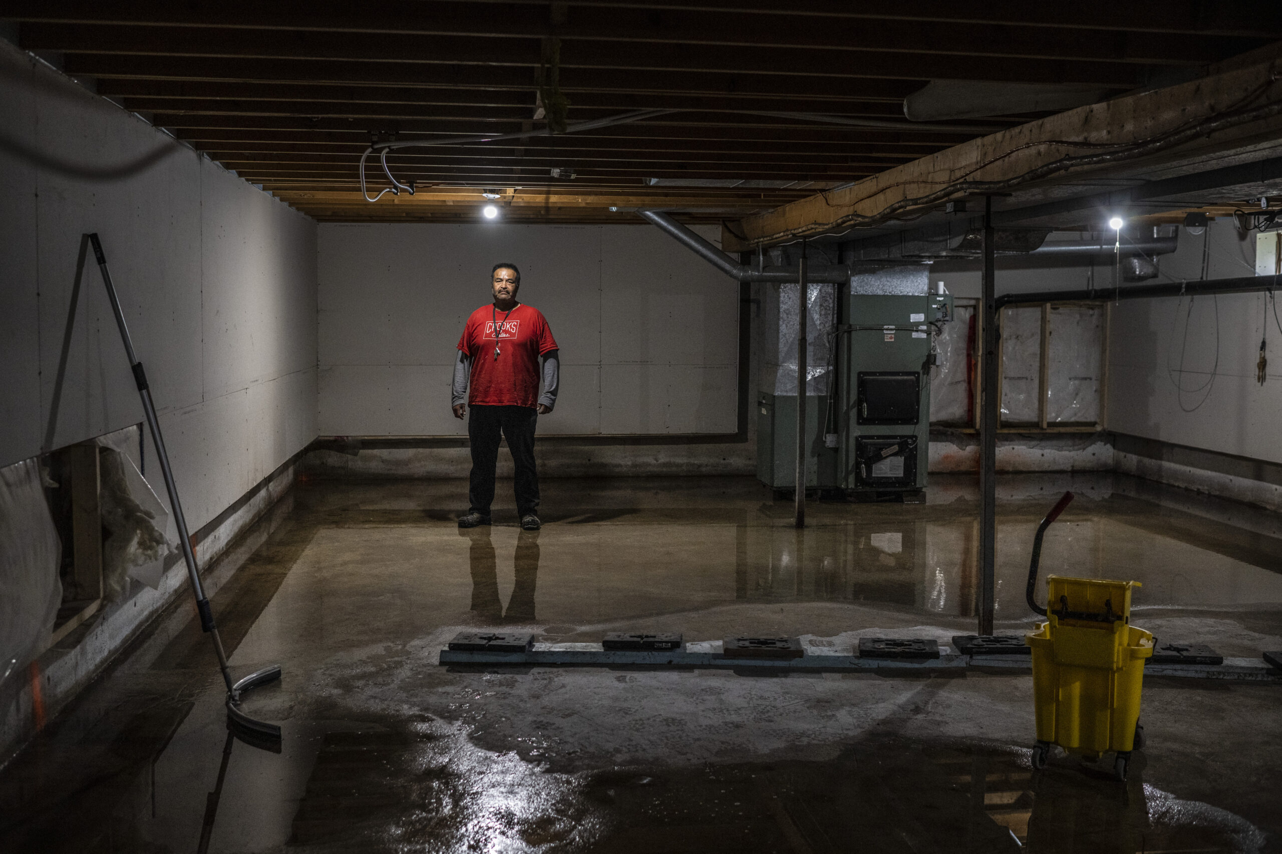 A man in a red shirt stands under a single lit bulb in a dark basement with water covering the floor. A long-armed, curved squeegee leans against the wall in the foreground