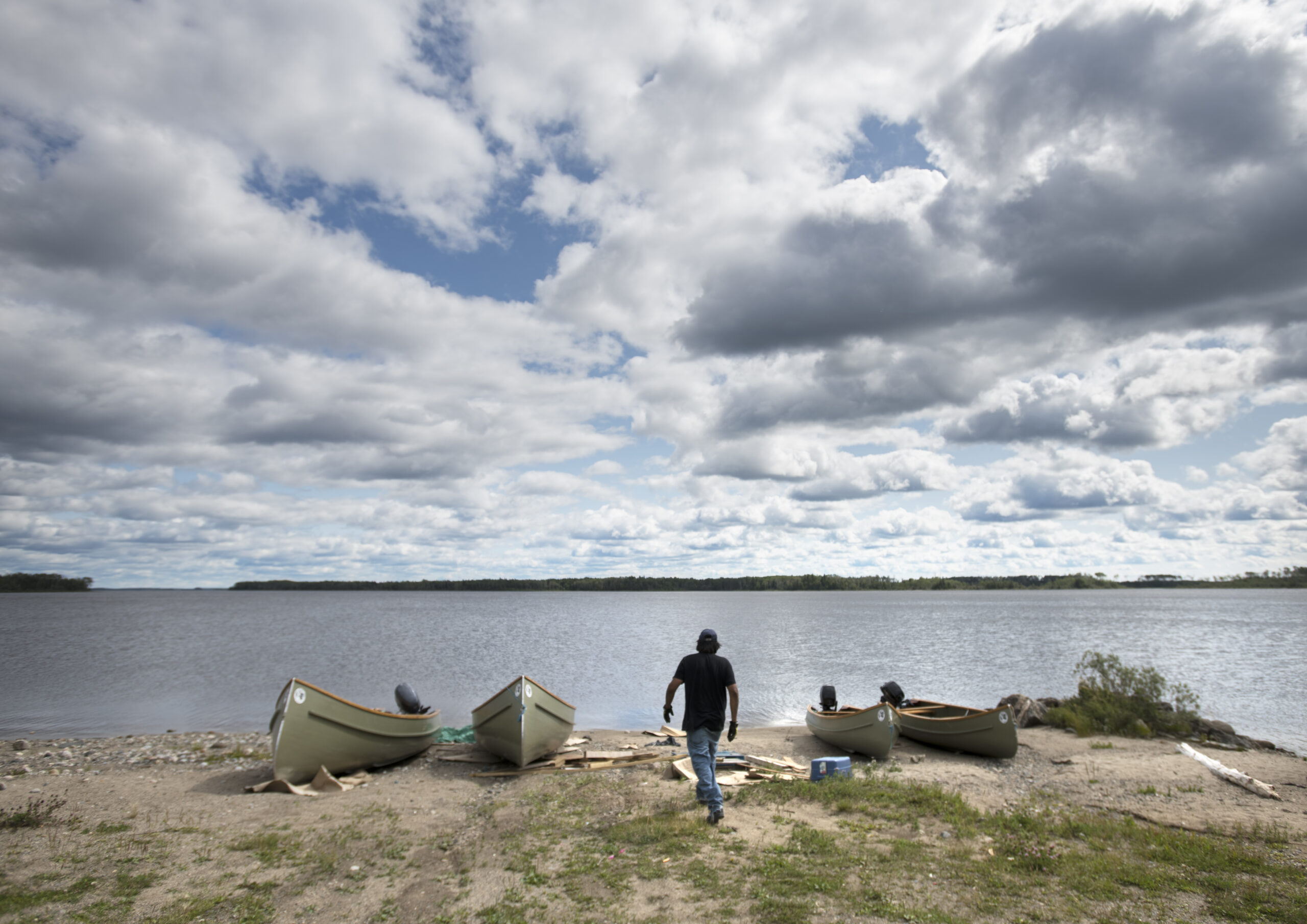 A man walks towards four canoes on the shore of a large lake on a cloudy day