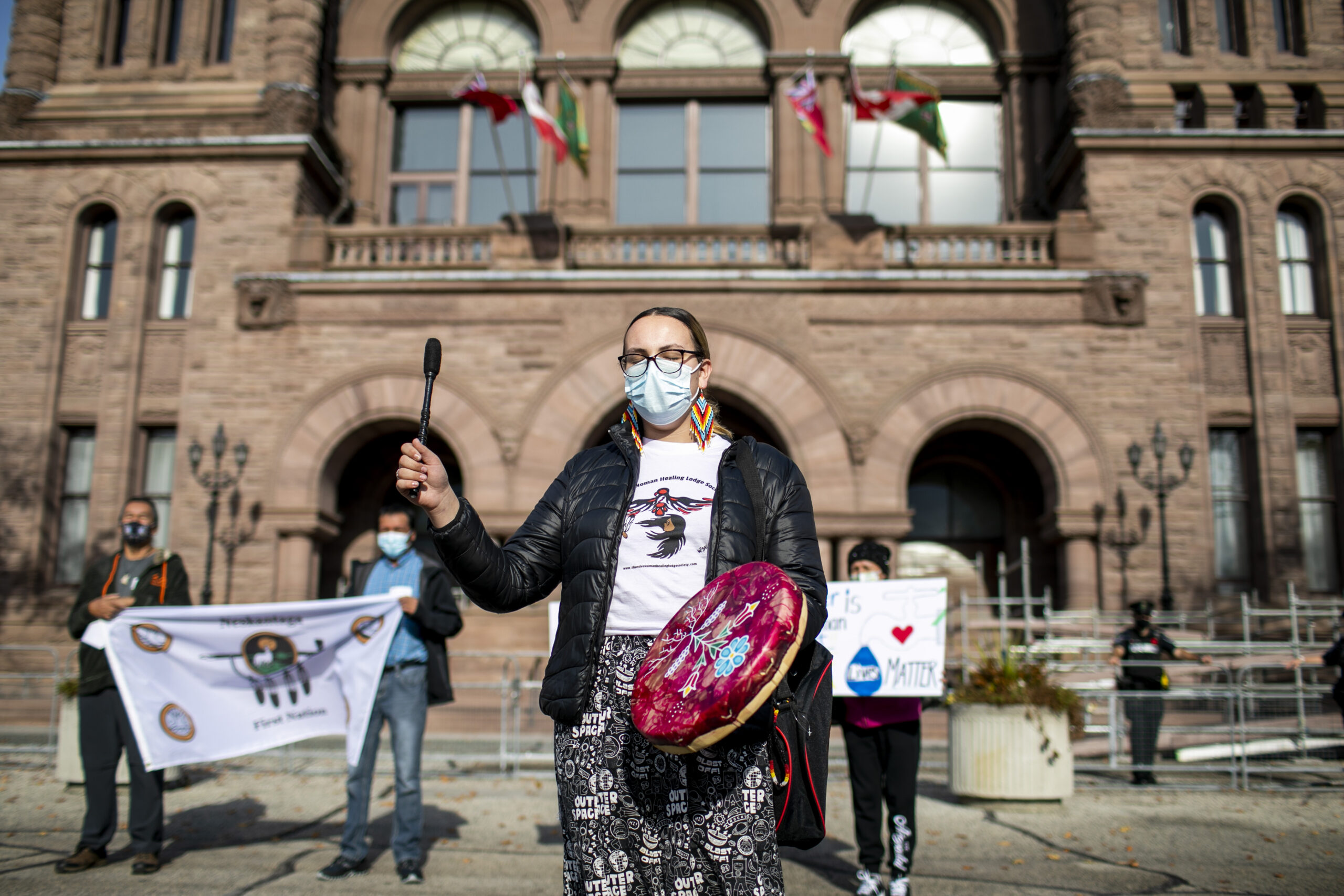 A woman in a medical face mask drums in front of Queen's Oark