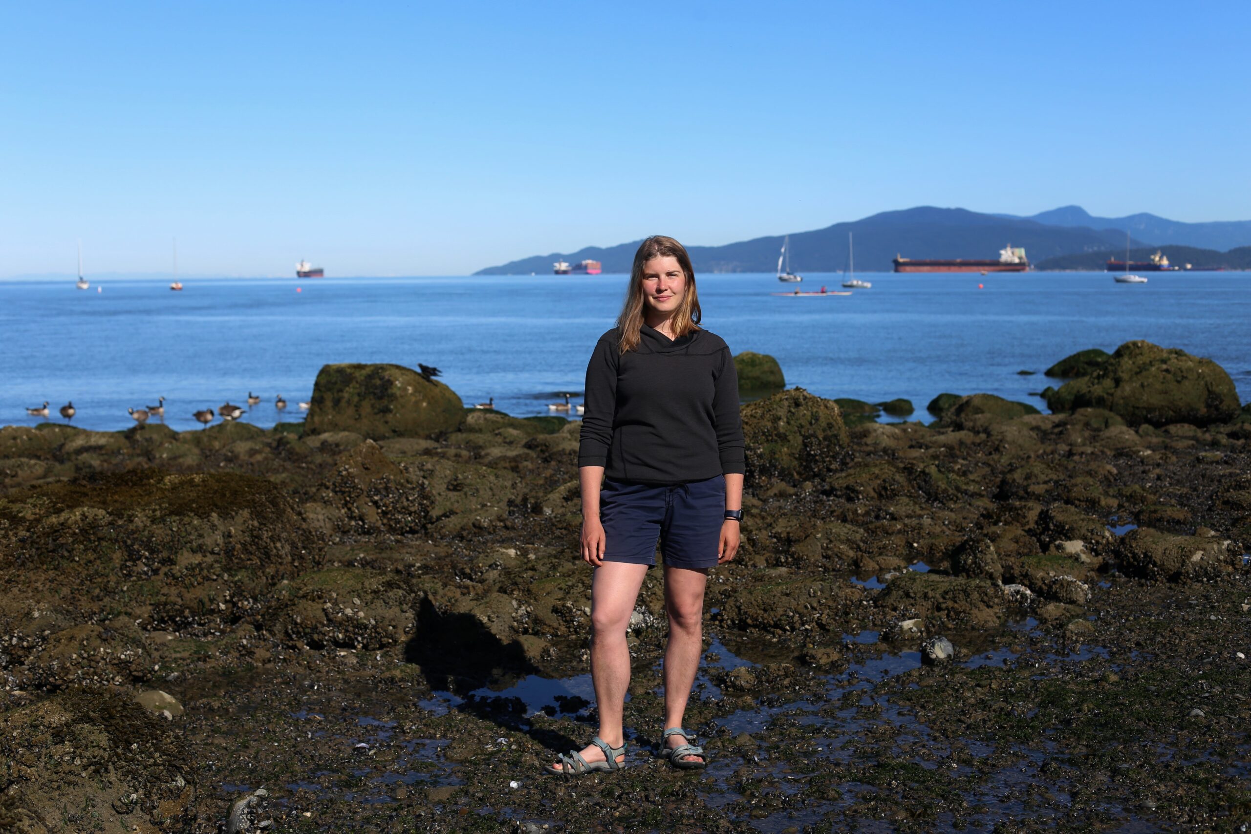 Amelia Hesketh, a research at the University of British Columbia stands in the intertidal zone at low tide on Kitsilano Beach with the ocean behind her and tanker ships visible in the distance.