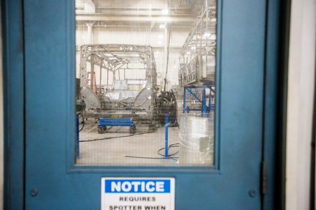 steel bus frames on wheeled carts are visible through the window of a blue door at New Flyer's Winnipeg facility
