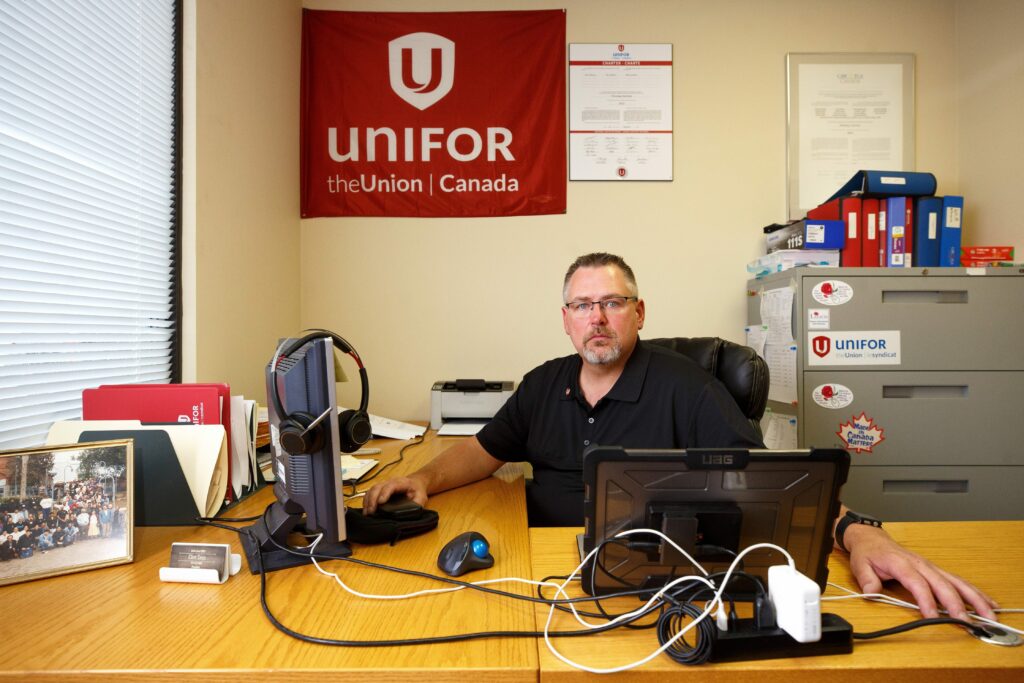 Union leader Clint Seys sits behind a desk in his office wearing a black polo shirt. A red Unifor flag hangs on the wall behind him.