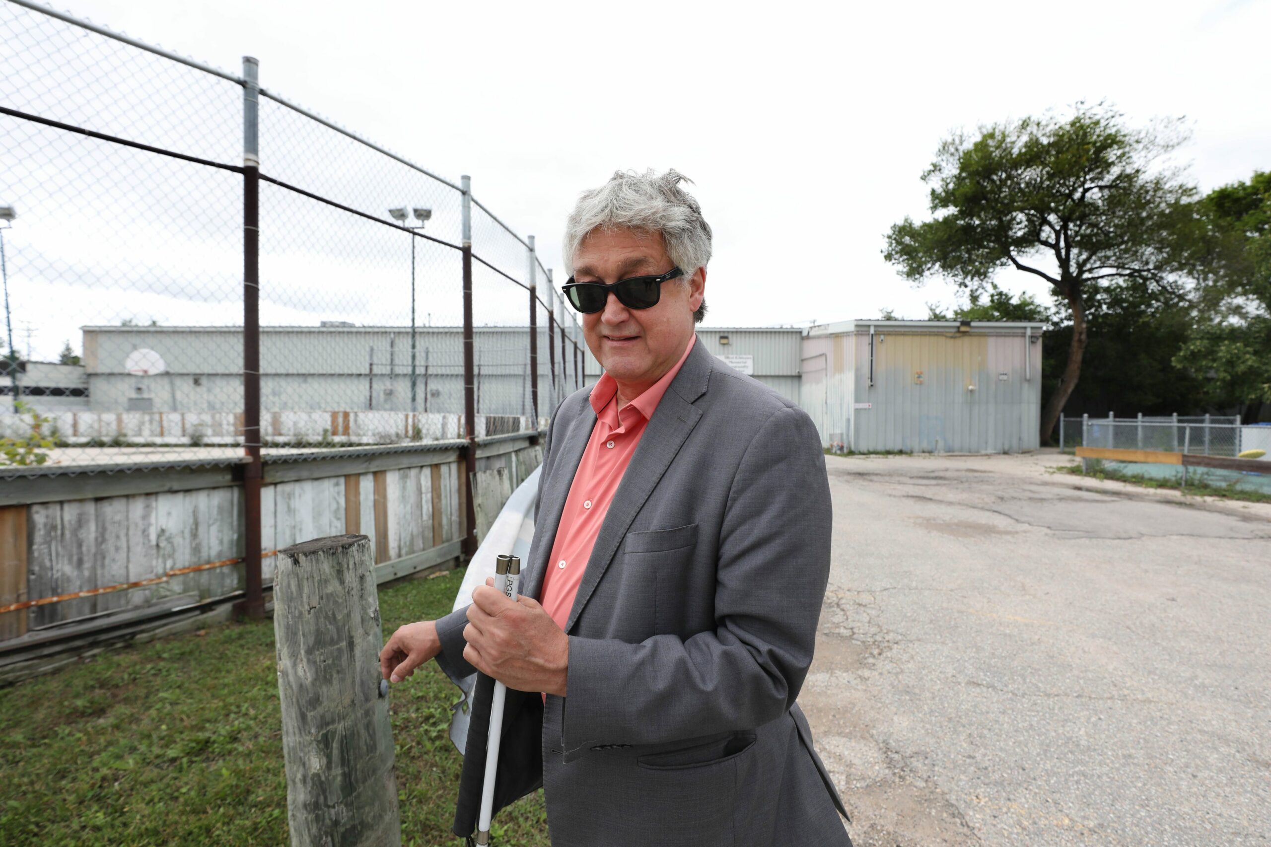 Ross Eadie, city councillor, stands outside the West Kildonan community centre wearing a grey jacket, salmon shirt and dark glasses. Behind him a high chain link fence encloses an outdoor arena