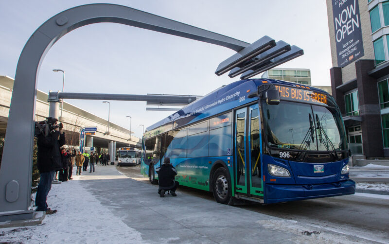 A blue batter-electric bus pulls into an overhead charging station at Winnipeg's airport while a small crowd watches from the sidewalk