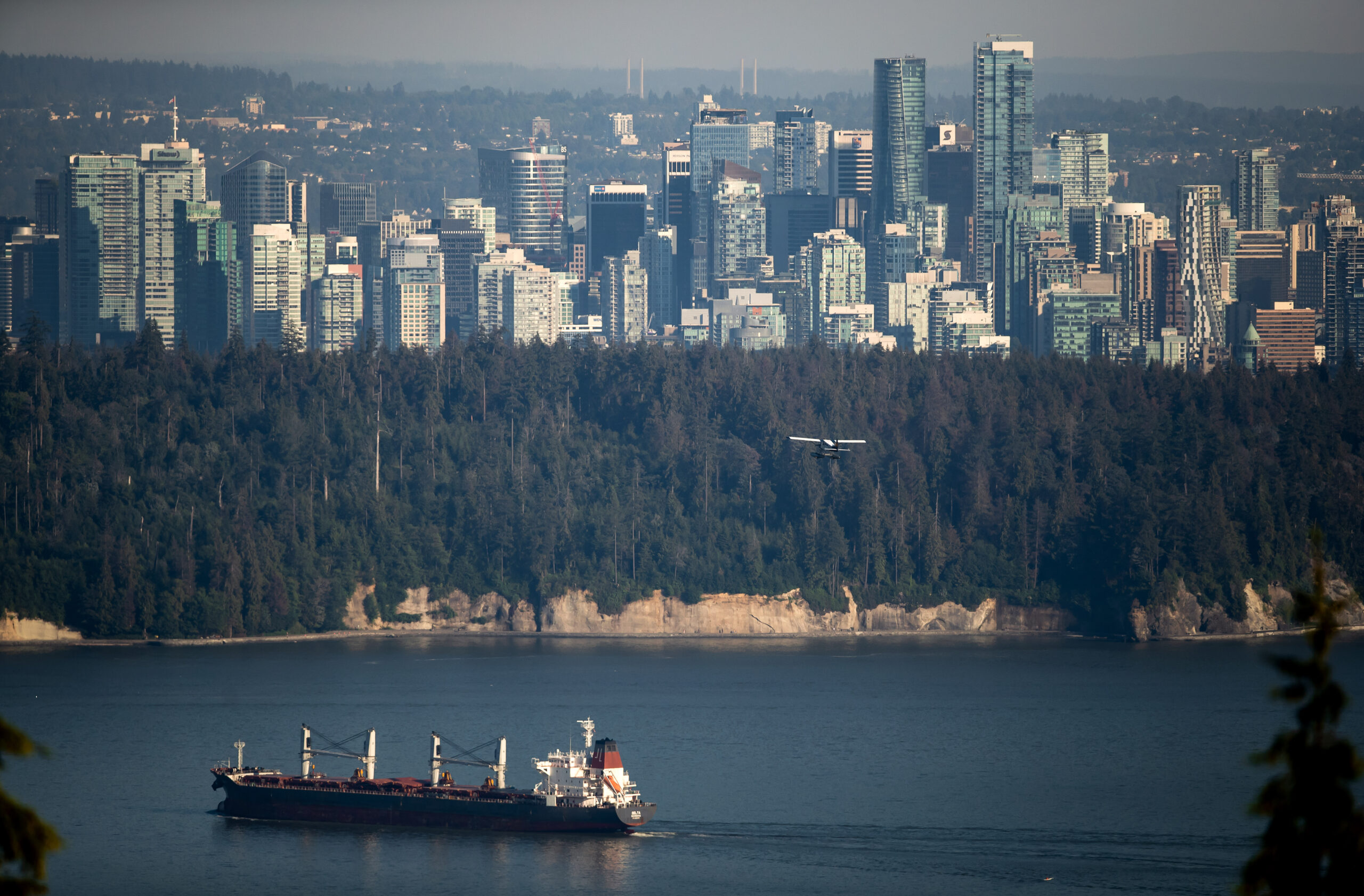 An ocean liner is seen in the foreground in a bay with Vancouver's skyline in the background and some trees in the middle of the photo. A small plane can also be seen.