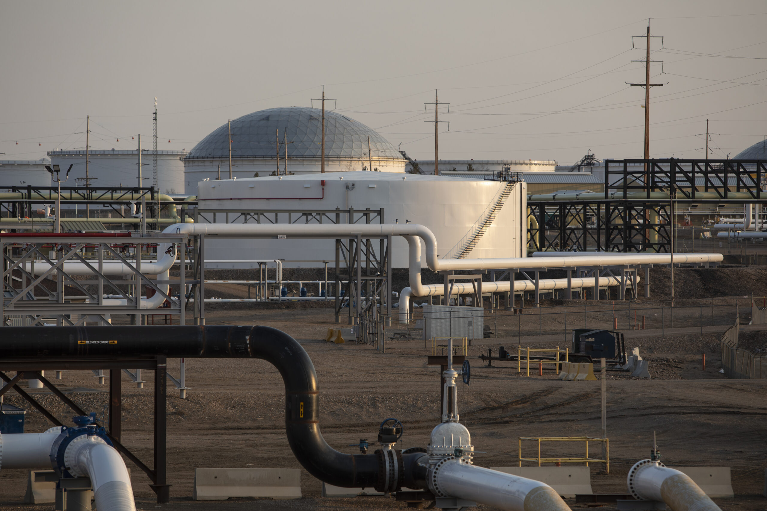 A series of pipes and storage tanks can be seen sitting atop a plain dirt field at dusk against a light brown sky.
