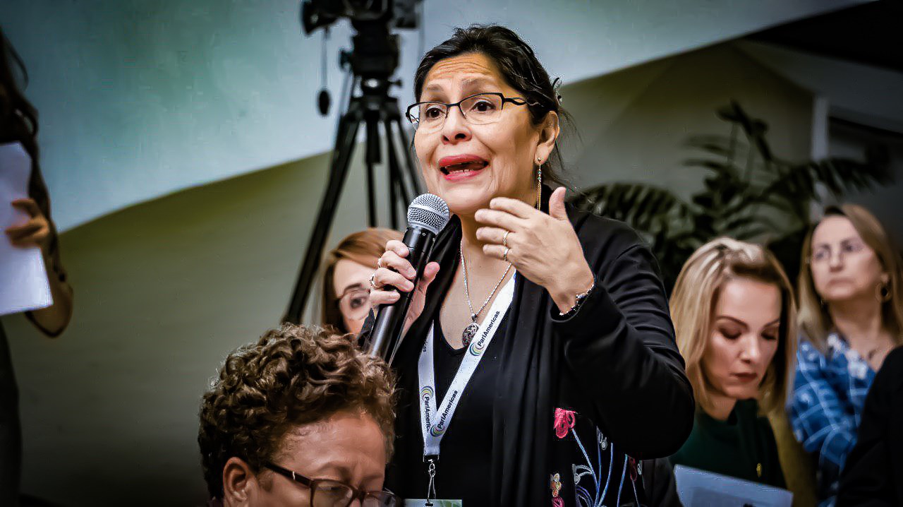 A woman wearing a lanyard speaks into a microphone while some people around her look down or to the side, perhaps reading something out of view.