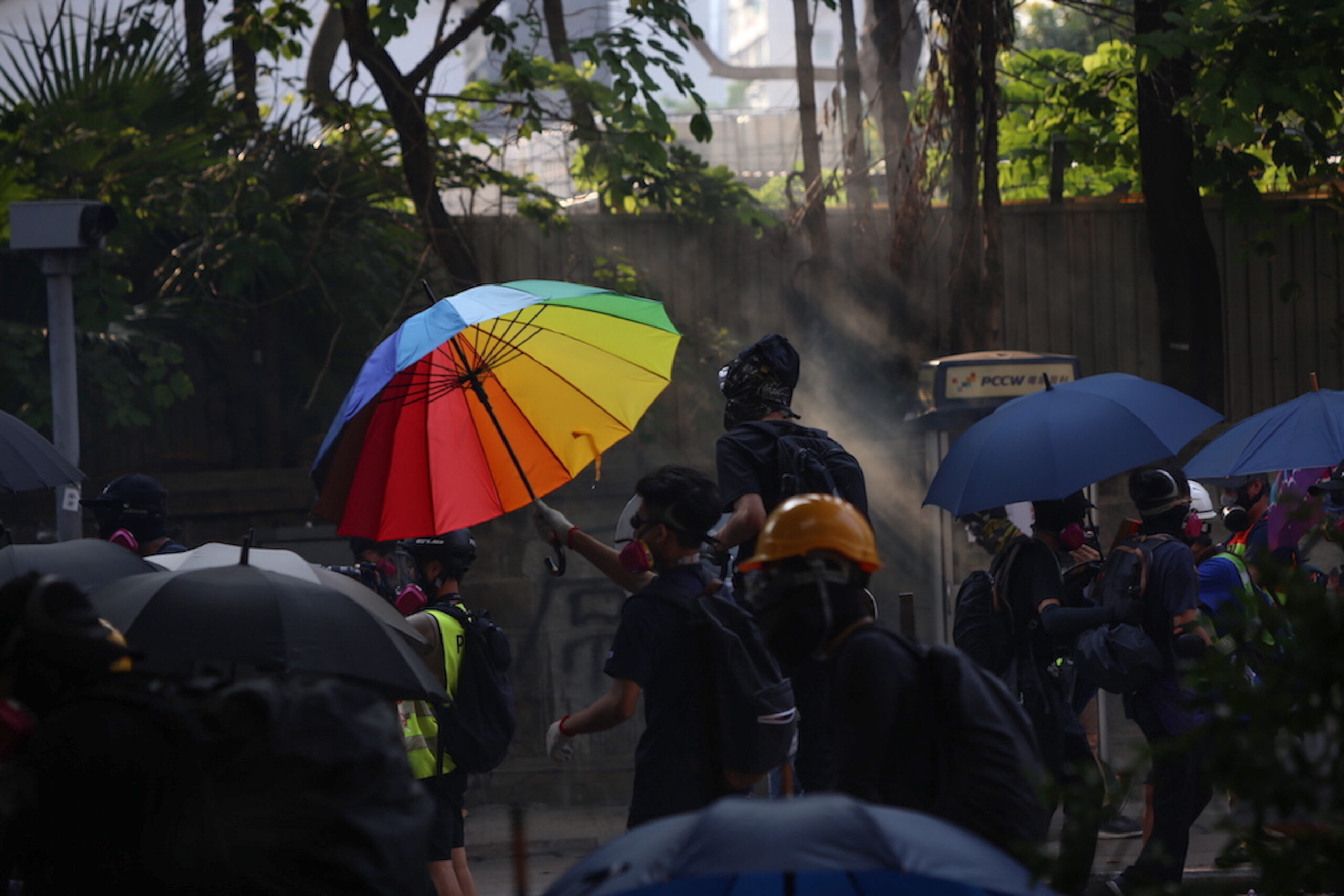 A protester clad in all-black clothing uses a bright rainbow umbrella as a shield against tear gas and surveillance by police.