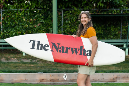 A member of The Narwhal holds a striped red and white surfboard with the logo