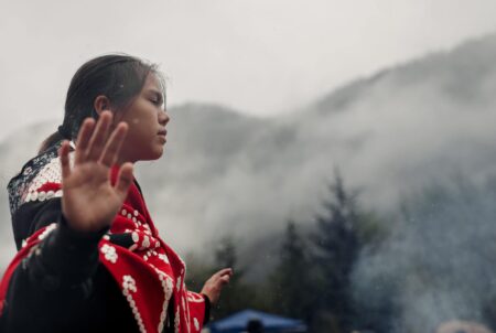 A Mamalilikulla youth dances in the rain at the nation's recently declared Indigenous Protected Area. She wears regalia, has her arms out and her eyes closed, and misty mountains are visible in the distance