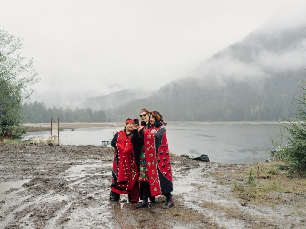 Mamalilikulla IPCA celebration three women in red and black regalia stand in front of the ocean and take a selfie, surrounded by mud, mist and rain